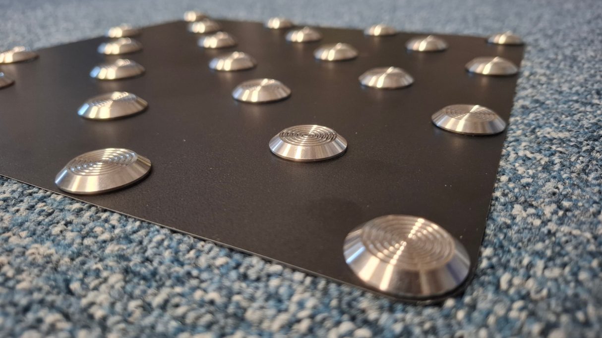 Schwarze Bodenplatte mit Metallnoppen auf blauer Teppichunterlage.